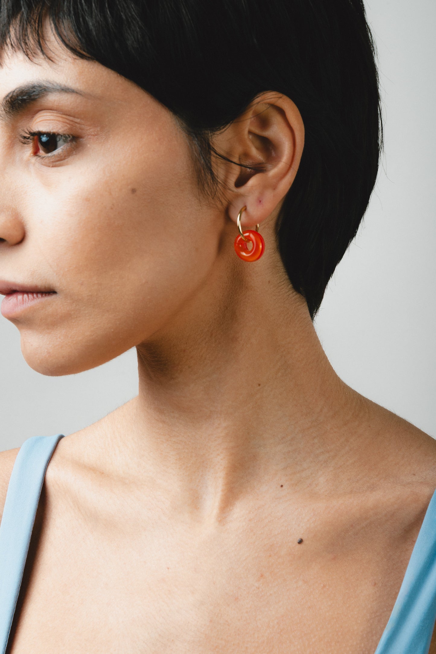 A pair of orange earrings in the shape of a donut, on a golden hoop, made from recycled glass