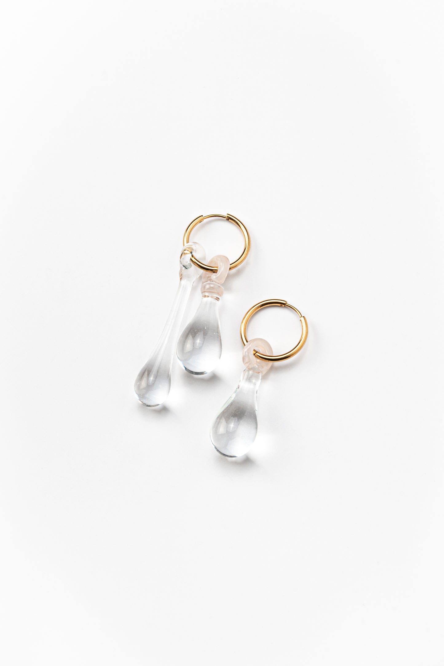Pair of assymetrical clear, transparent drop-shaped earrings on a golden hoop. made from recycled glass