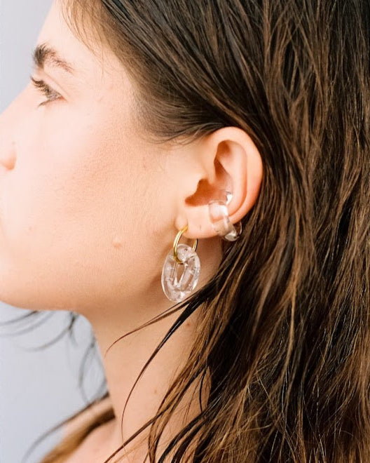 A pair of clear, transparent oval earrings on a golden hoop, made from recycled glass