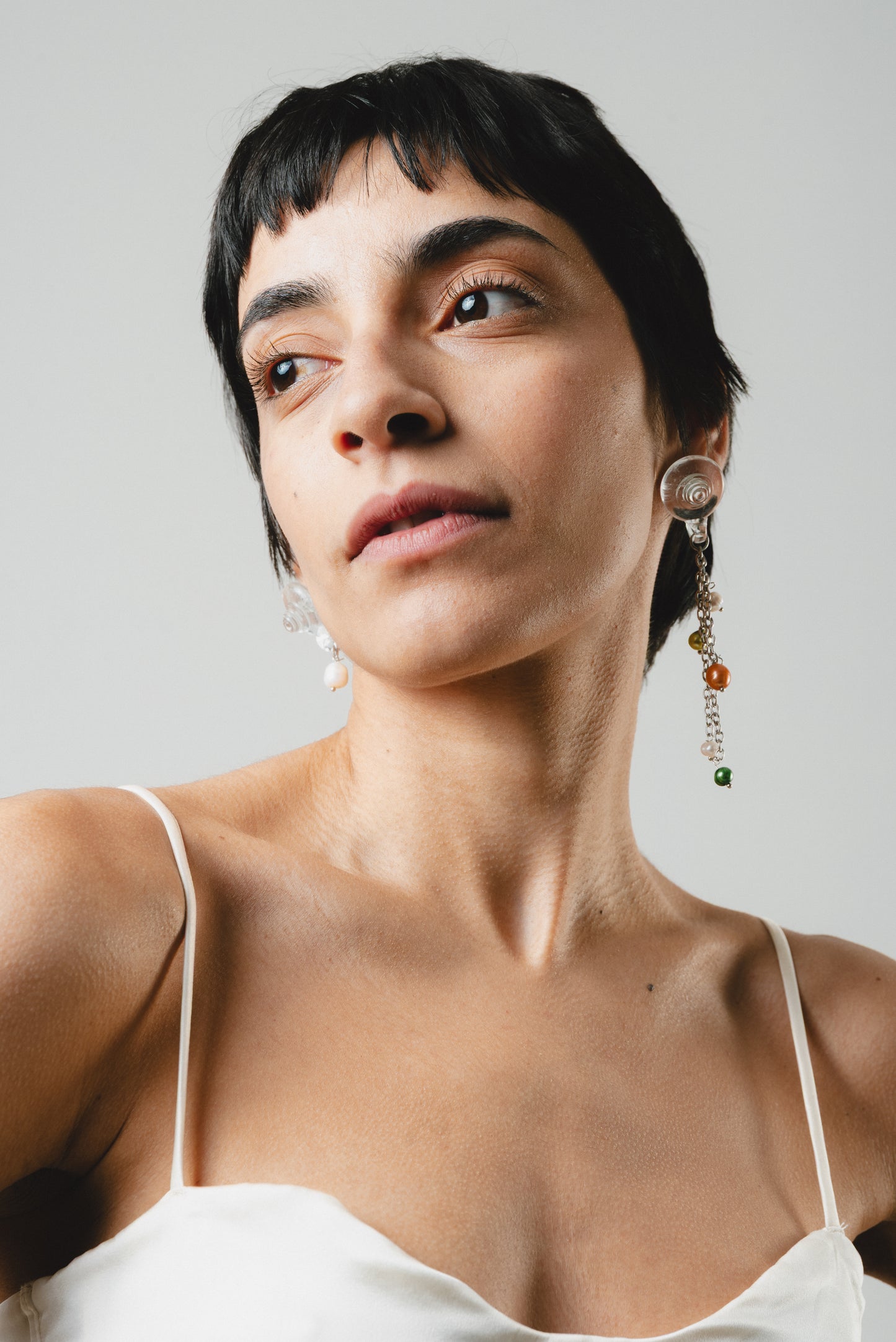 Drop earrings with clear shell-shaped studs made of recycled glass. Each earring has a silver chain hanging down, with small round pearls in green, orange, and white attached at different points along the chain.