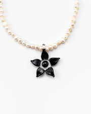Necklace made of small white river pearls, featuring a gold toggle clasp and a large black flower pendant made from recycled glass