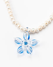 Necklace made of small white river pearls, featuring a gold toggle clasp and a large light blue flower pendant made from recycled glass