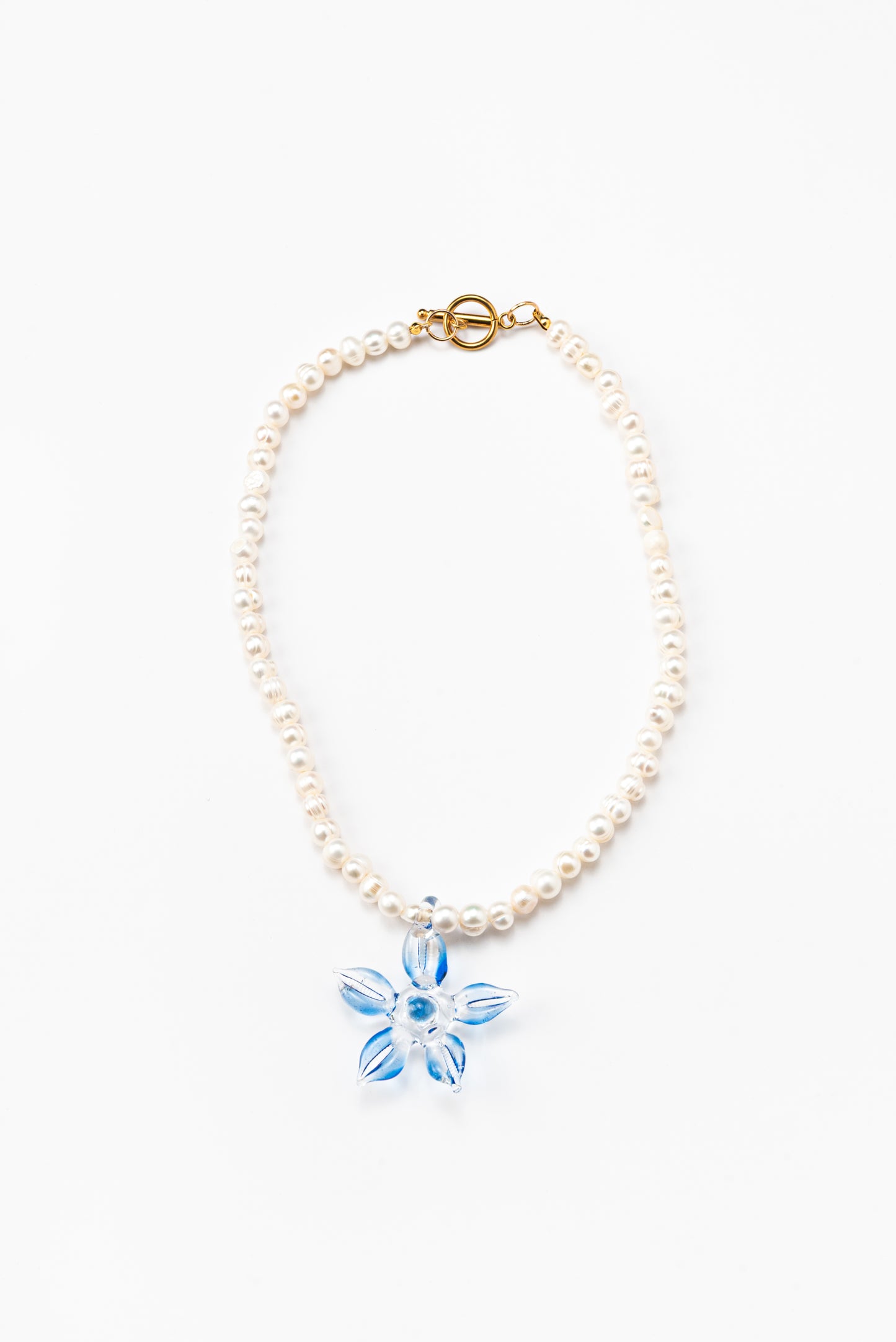 Necklace made of small white river pearls, featuring a gold toggle clasp and a large light blue flower pendant made from recycled glass