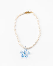 Necklace made of small white river pearls, featuring a gold toggle clasp and a large light blue flower pendant made from recycled glass