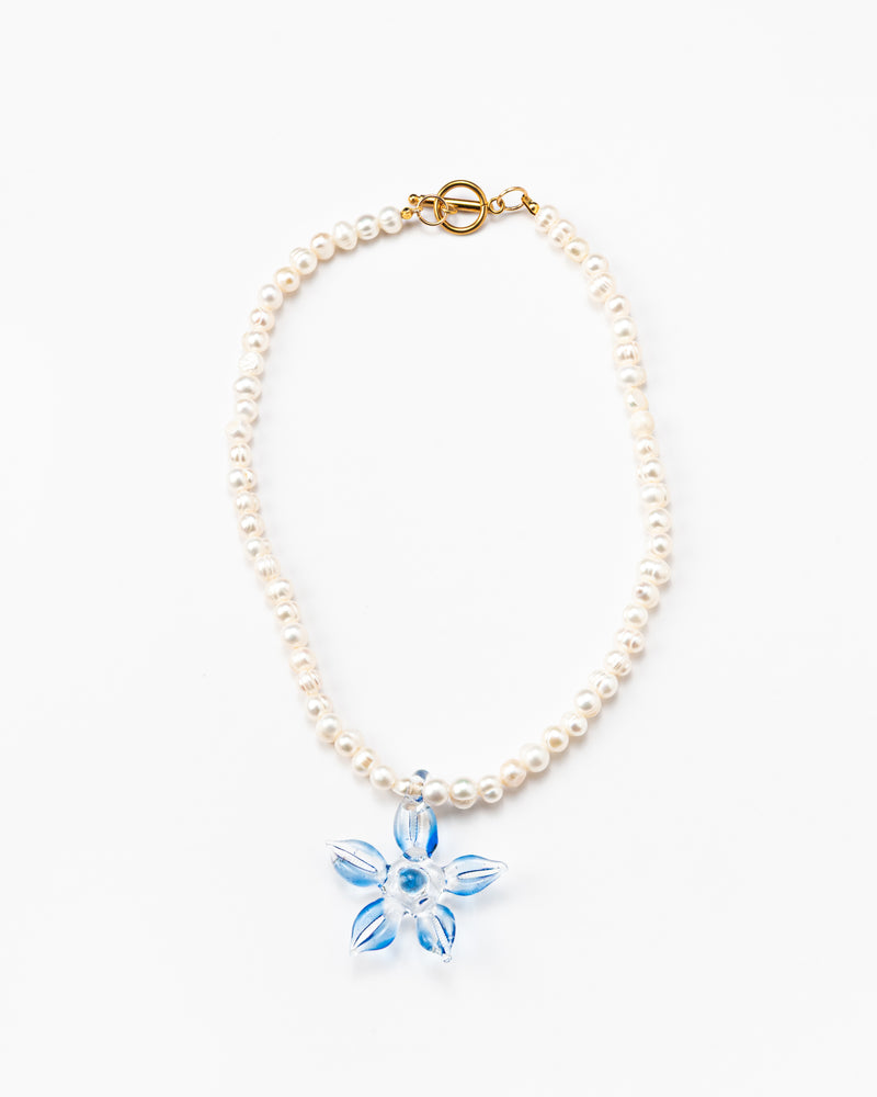 Necklace made of small white river pearls, featuring a gold toggle clasp and a large light blue flower pendant made from recycled glass