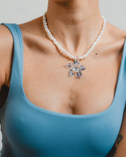 Necklace made of small white river pearls, featuring a gold toggle clasp and a large light blue flower pendant made from recycled glass