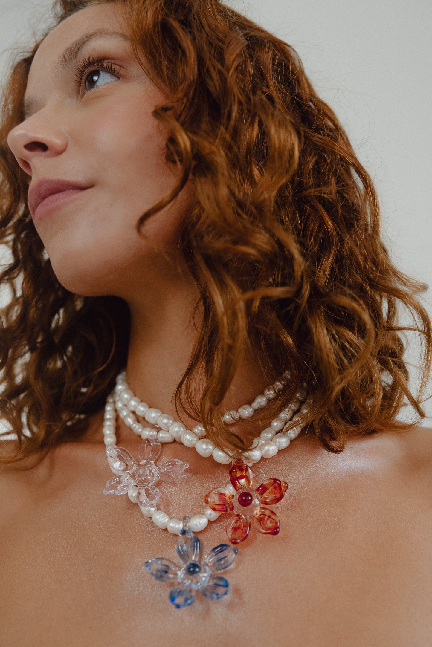 Pimpollo necklaces made of small white river pearls, featuring a gold toggle clasp and large light blue, pink, and red flower pendants made from recycled glass