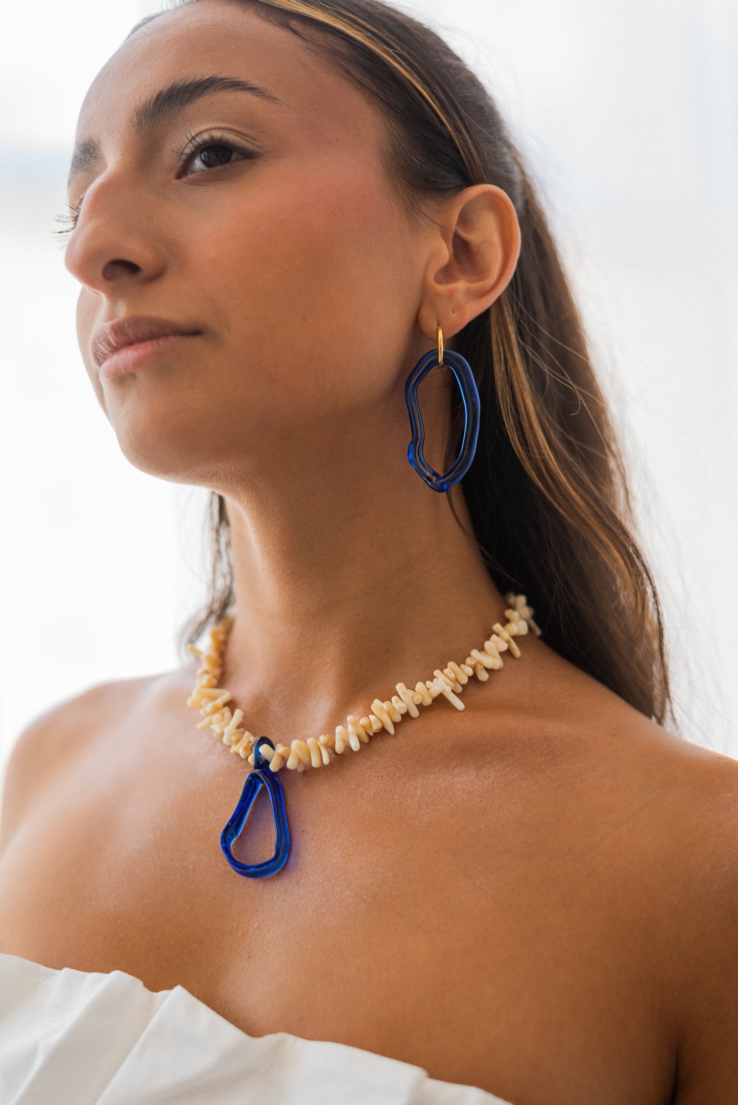 Necklace made of beige sea shell beads with a loop-shaped blue recycled glass pendant and a gold toggle clasp