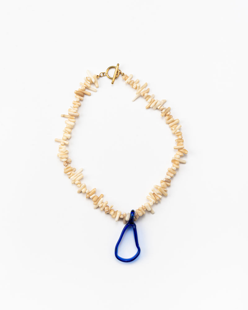 A necklace made of beige sea shell beads with a loop-shaped blue recycled glass pendant and a gold toggle clasp