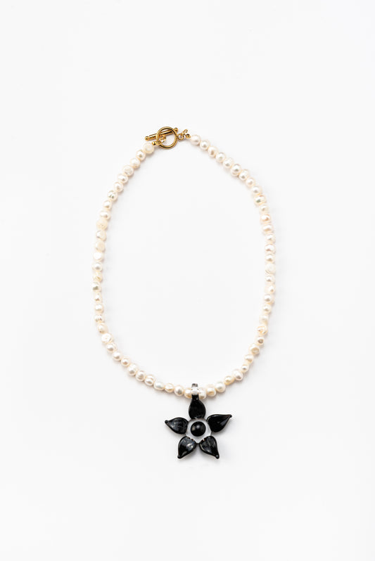Necklace made of small white river pearls, featuring a gold toggle clasp and a large black flower pendant made from recycled glass