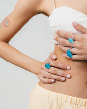 The model is wearing two types of rings: clear ring with a turquoise textured glass detail on top, made from recycled glass; and a clear ring with a turquoise sphere on top, made from recycled glass